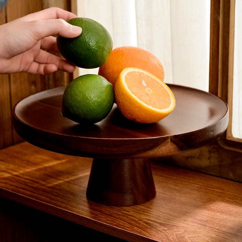 A hand places limes and an orange on a wooden pedestal stand, with a rustic wooden background and soft light coming through white curtains.