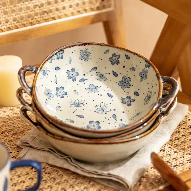 A stack of three ceramic bowls with blue floral patterns on the inside. They are placed on a folded napkin, with a woven chair and a candle visible in the background. The setting is warm and rustic.