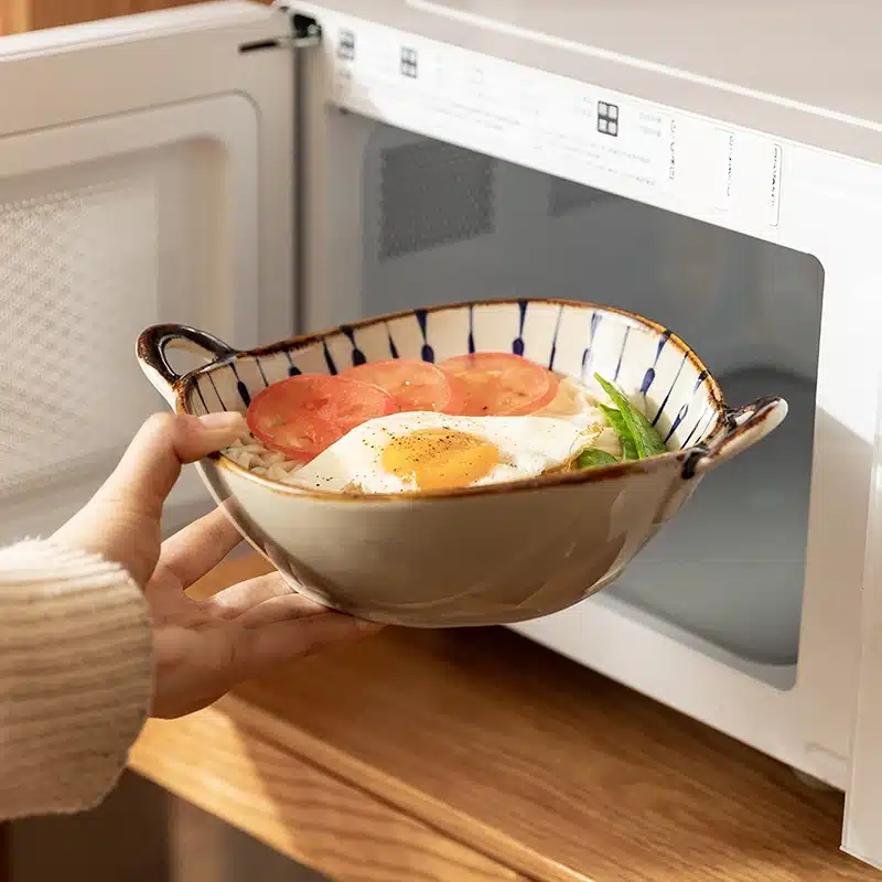 A person holding a ceramic bowl with a fried egg, sliced tomatoes, and greens, placing it in a microwave. The microwave door is open, and the scene is set on a wooden surface.