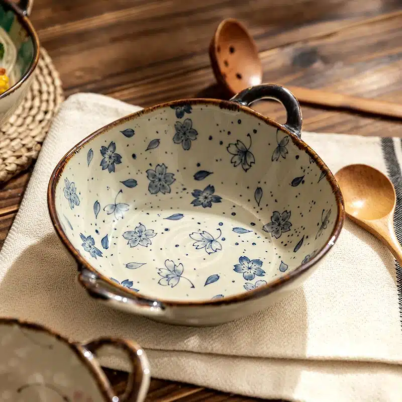 A ceramic bowl with blue floral patterns sits on a beige cloth on a wooden table. Two wooden spoons are nearby. The bowl has two small handles on each side.