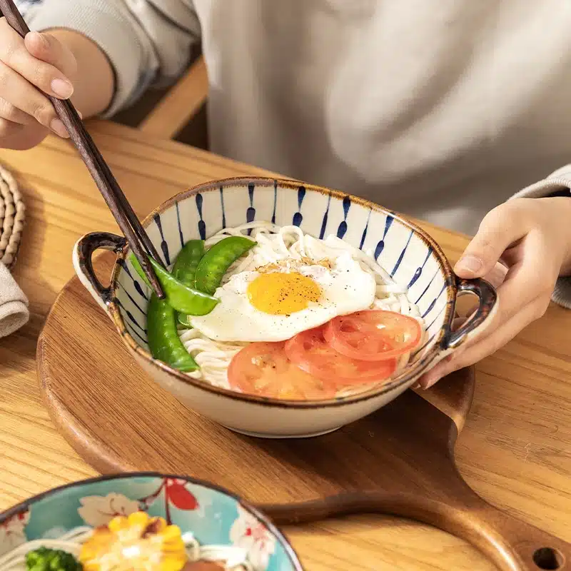 A person holding chopsticks is about to eat a bowl of noodles topped with a fried egg, sliced tomatoes, and green vegetables. The bowl is placed on a wooden board.