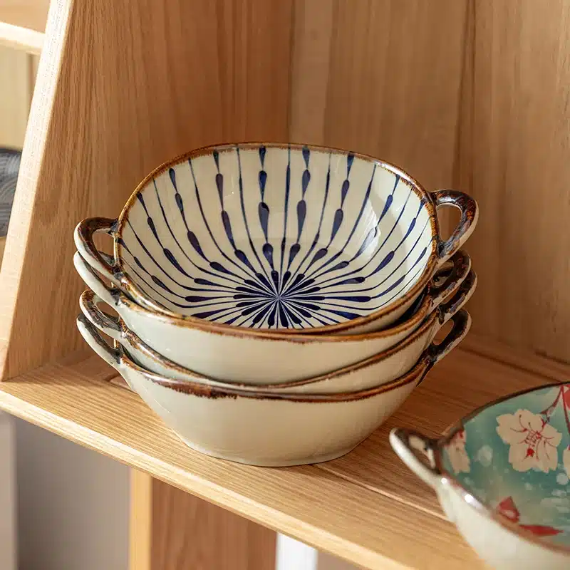 Stack of ceramic bowls with blue radial patterns on a wooden shelf. One bowl to the right features a floral design on a turquoise background. The shelf is part of a light wood display unit.