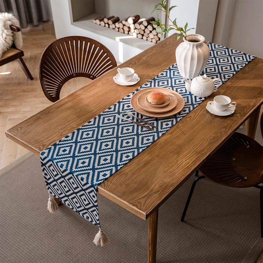 A wooden dining table with a blue and white geometric-patterned runner. It is set with two white teacups, a plate with a pastry, and a white vase with branches. Two wooden chairs are positioned on either side.