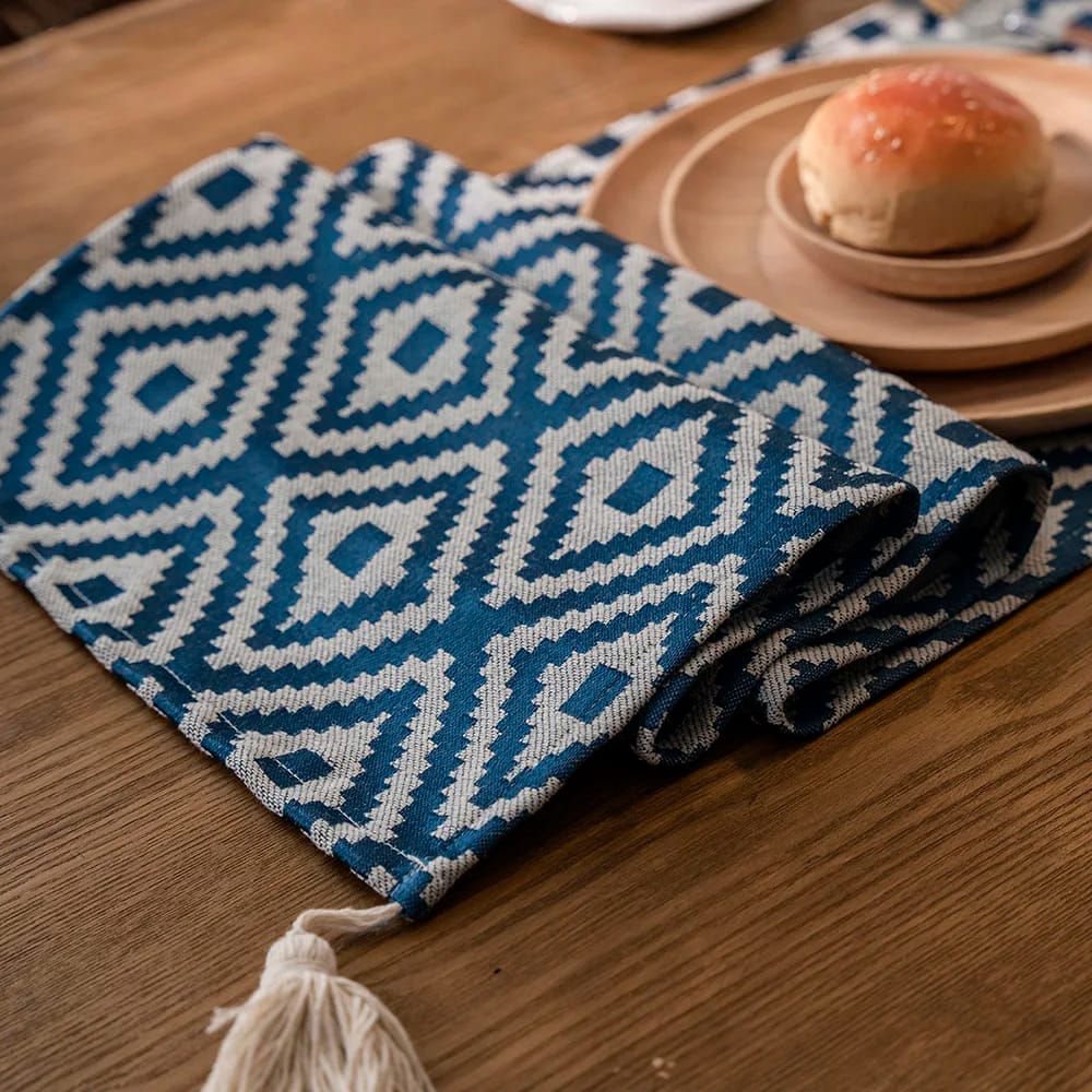 A wooden table with a blue and white geometric-patterned table runner and a round wooden tray holding a small bread roll. A white tassel hangs from the end of the runner.