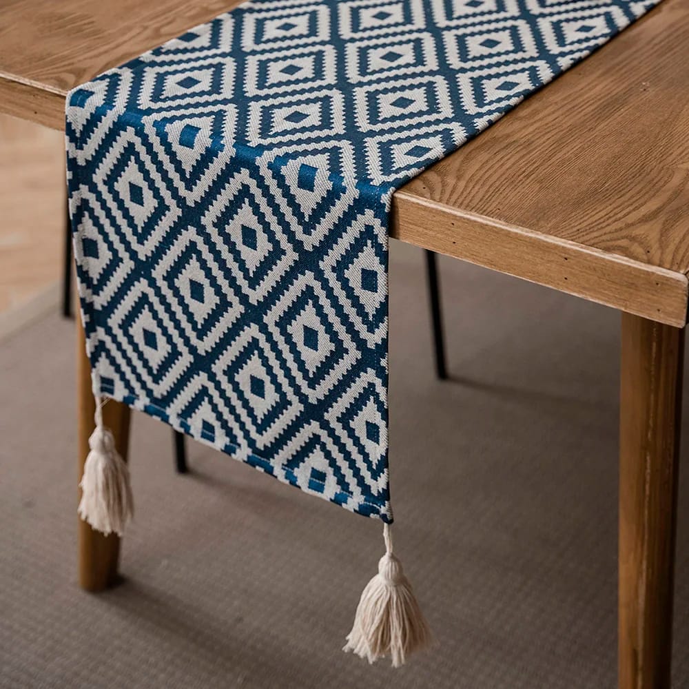 A wooden table is adorned with a blue and white geometric patterned table runner featuring diamond shapes. The ends of the runner are decorated with white tassels, and the table rests on a light brown carpet.