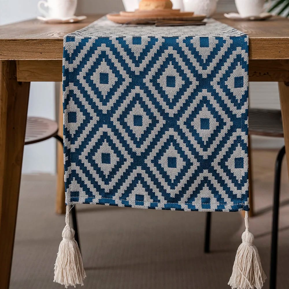 A close-up of a wooden dining table decorated with a blue and white geometric-patterned table runner featuring tassels. In the background, there are two chairs and white teacups on the table.