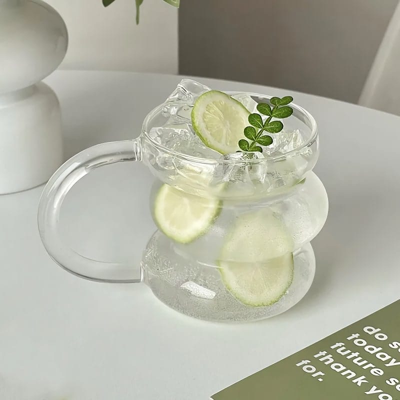 A unique, wavy glass mug filled with ice water, lemon slices, and a small green leaf sits on a white surface. A white lamp is partially visible in the background, with part of a green and white card in the foreground.