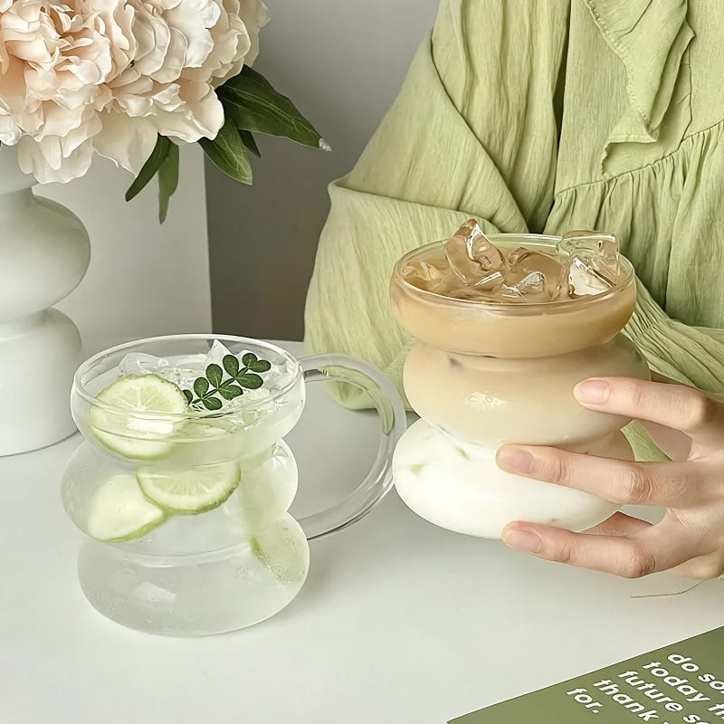 A person wearing a light green outfit holds a double-walled glass filled with iced coffee. Beside it, there is another glass containing a drink with lime slices and a sprig of greenery. A vase with white flowers is in the background.