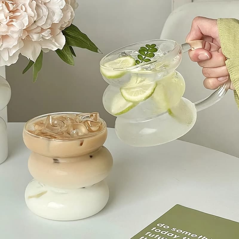 A hand pours lemon and mint water from a uniquely shaped glass jug into a matching glass. Another similar glass, filled with iced coffee, sits on a table next to flowers and a green card with white text.