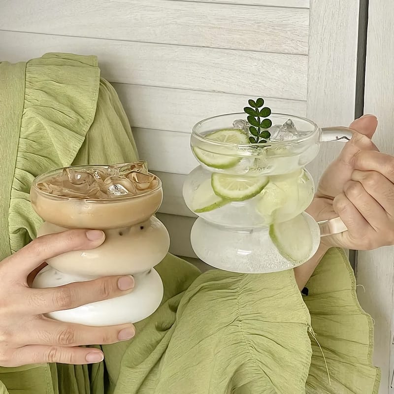 A person in a green outfit holds two uniquely shaped mugs. One mug contains iced coffee with milk and ice cubes, and the other has a clear drink with ice, cucumber slices, lime slices, and a garnish.