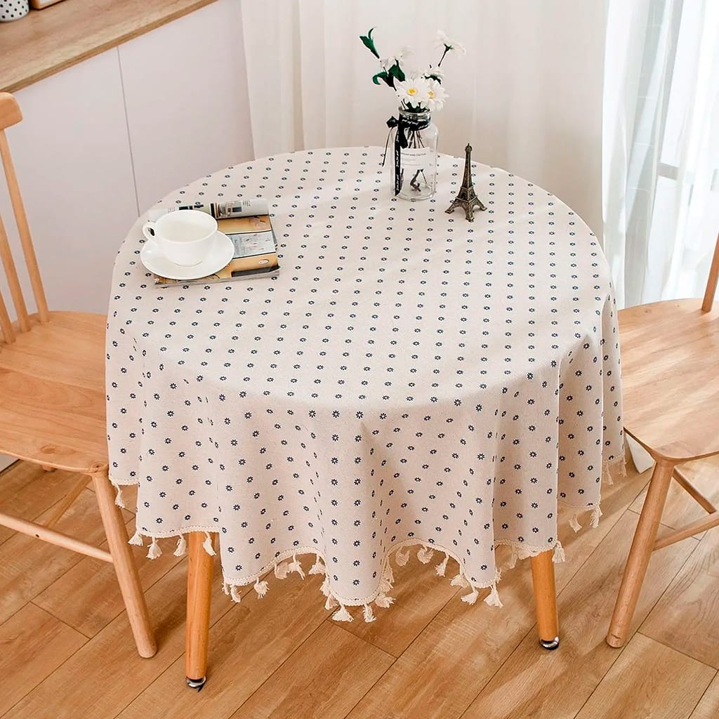 A small round wooden table with a beige tablecloth featuring small blue polka dots stands between two wooden chairs. On the table are a coffee cup, a book, a small Eiffel Tower figurine, and a vase with flowers. A curtain filters natural light.