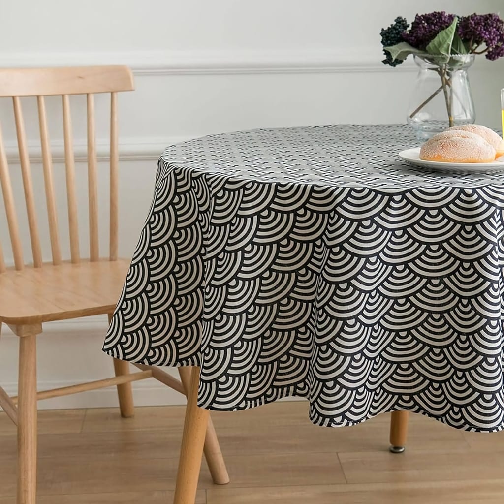 A wooden chair and a round table with a black and white geometric-patterned tablecloth. A glass vase with purple flowers and a plate with a bread roll are on the table. The setting is on a wooden floor with a white wall backdrop.