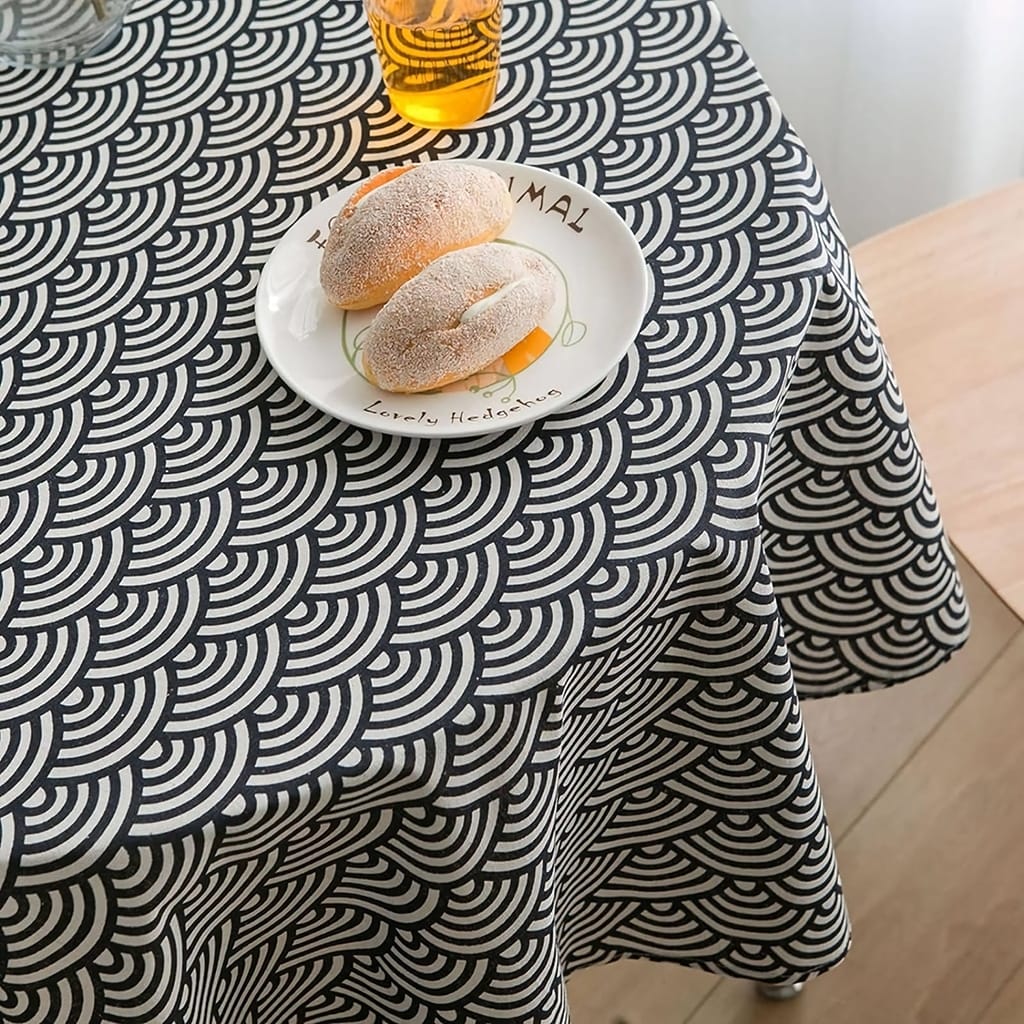 A table with a black and white geometric patterned tablecloth. On the table, theres a plate with two pastries and a glass of orange juice. The table is next to a window with natural light coming in.