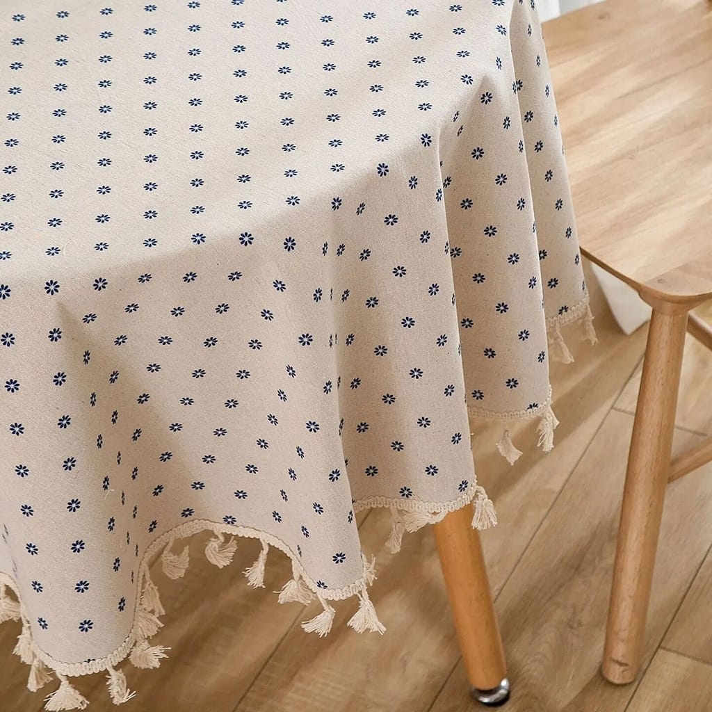 A wooden table with a patterned tablecloth featuring small blue flowers and fringe edges. The table is positioned on a wooden floor next to a matching wooden chair.