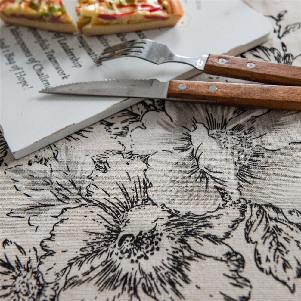 Tablecloth with black floral patterns beneath a white plate with partially eaten pizza slices. Two wooden-handled utensils, a fork and a knife, are placed beside the plate.