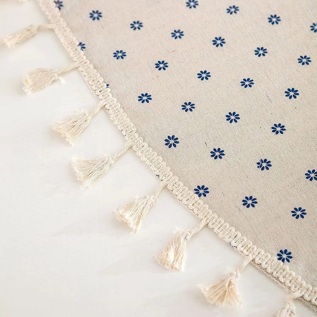 Close-up of a beige fabric with small blue floral patterns and a fringe of tassels along the edge, placed on a smooth white surface.