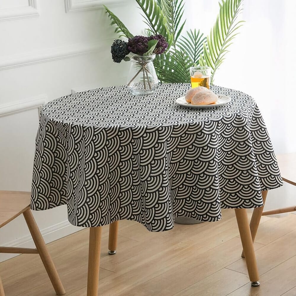 A round table with a black and white patterned tablecloth. On top, theres a vase with flowers, a glass of orange juice, and a plate with a piece of bread. Sunlight brightens the room, and green plants are in the background.