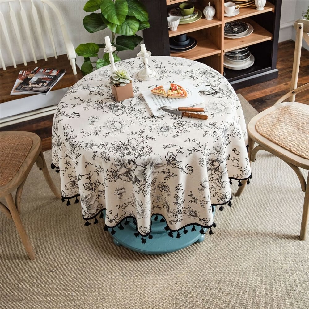 A cozy dining area with a round table covered by a floral-patterned tablecloth. The table has two candles, a succulent, and a plate with pizza slices. Two wooden chairs with woven seats are around the table. A shelf and plant are in the background.