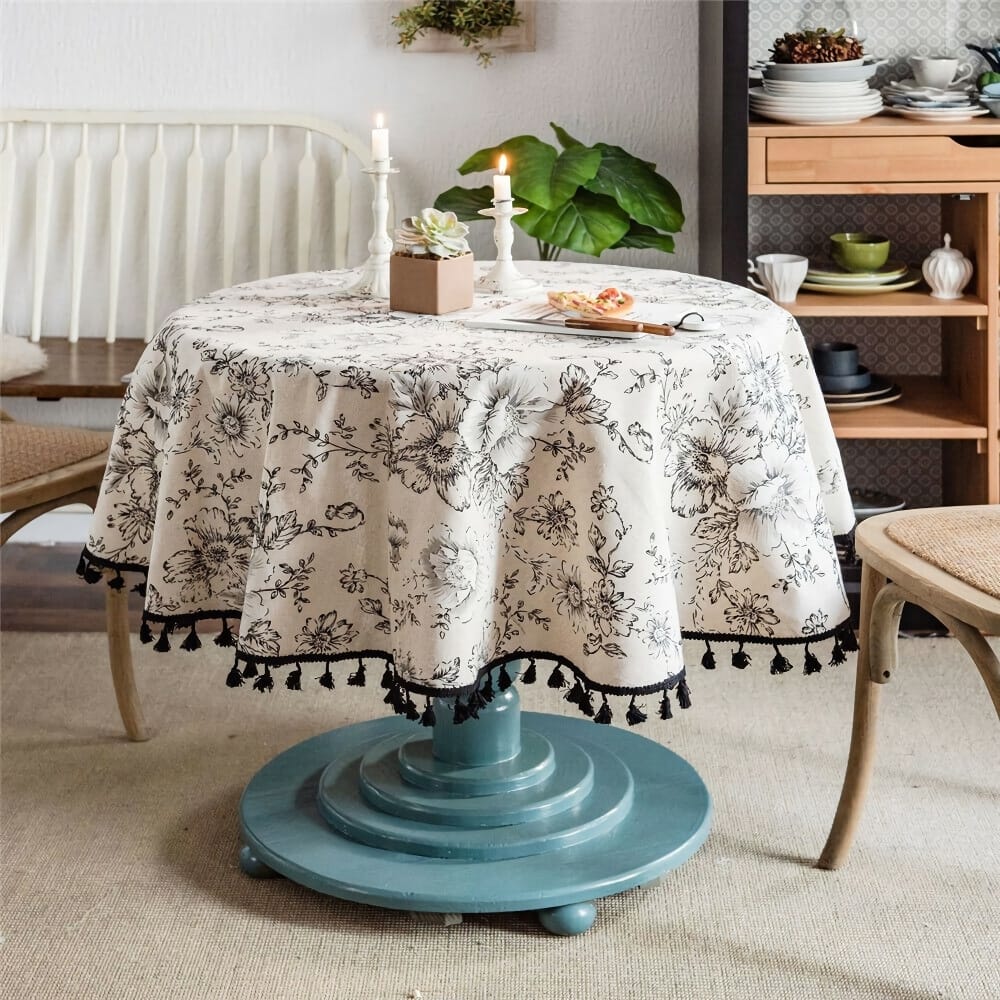 A cozy dining area with a round table covered in a floral-patterned cloth with tassels. Two lit candles and a small plant decorate the table. Surrounding chairs and a shelf with dishes are visible in the background.