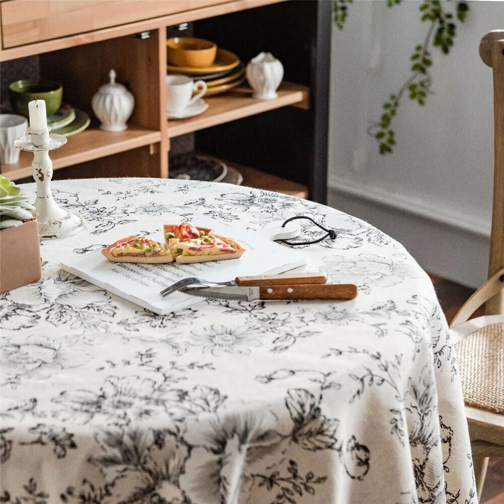 A round table with a floral tablecloth displays a cutting board with sliced pizza. Nearby are a fork and knife with wooden handles, a candle in a holder, and a cardboard pizza box. A wooden shelf in the background holds dishes and plants.