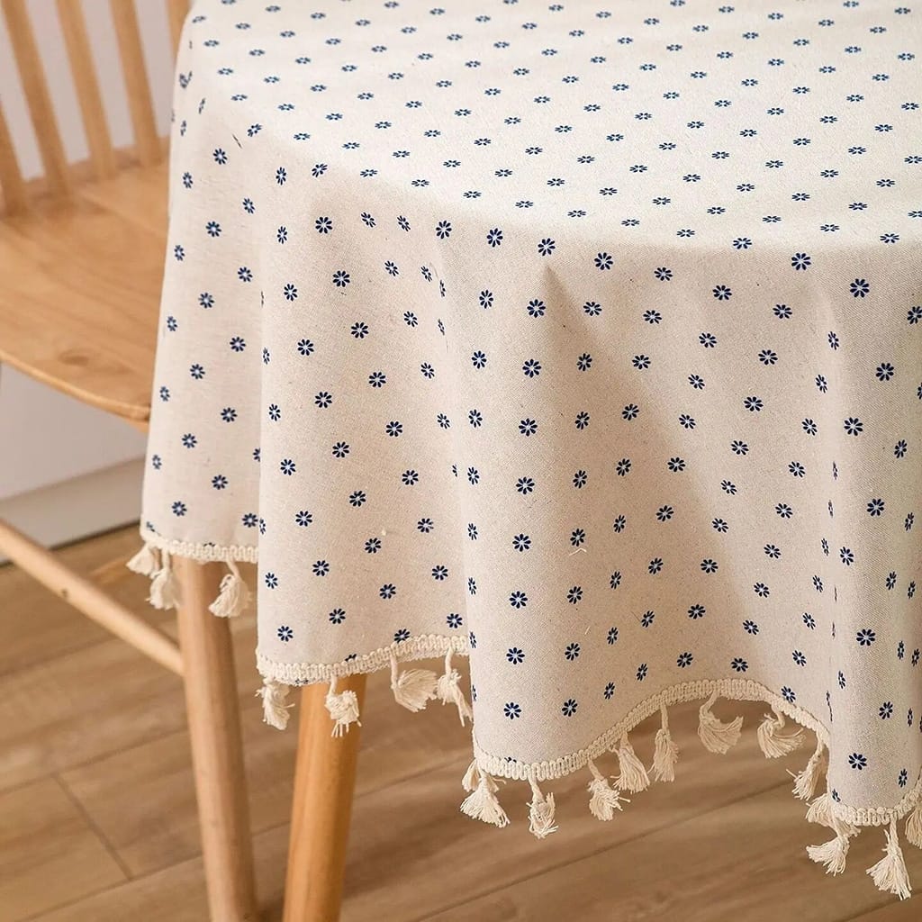 A round wooden table covered with a beige tablecloth featuring a pattern of small blue flowers. The tablecloth has tassels along the edge. There is a wooden chair next to the table on a wooden floor.