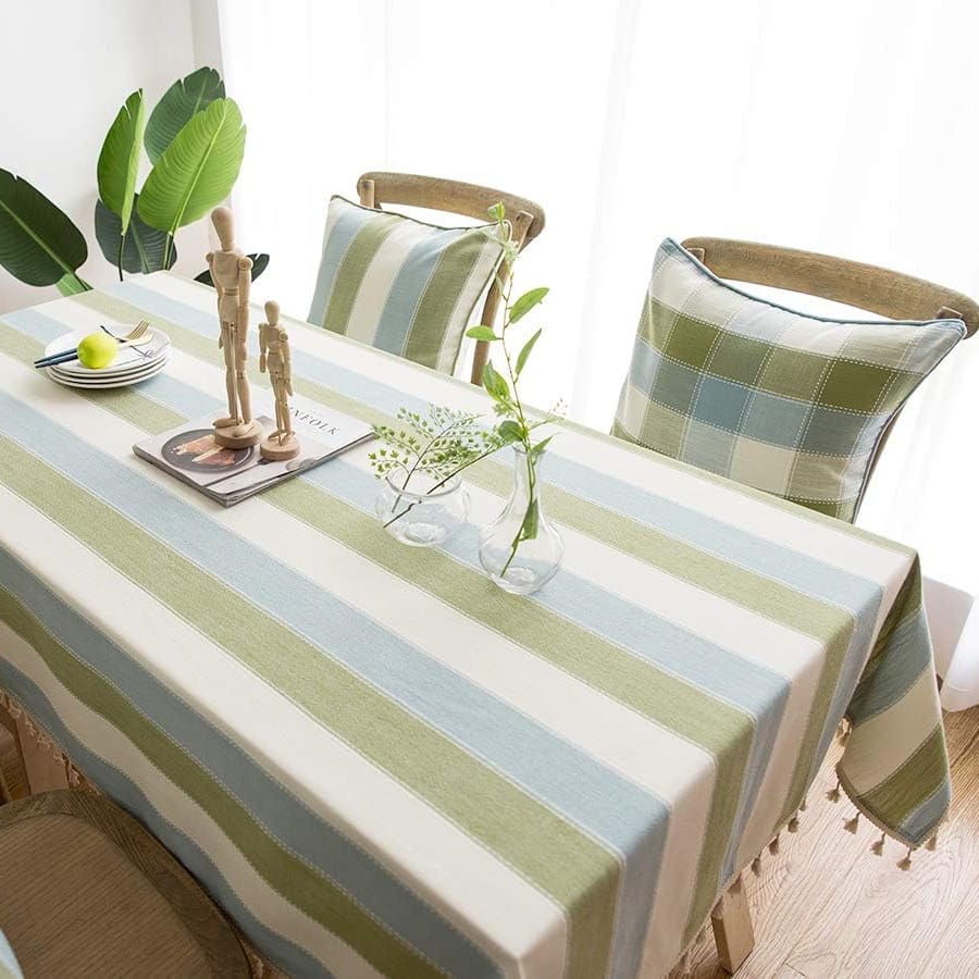 A dining table set with a striped tablecloth featuring blue, white, and green stripes. It includes neutral-colored wooden chairs with checkered cushions, a decorative wooden sculpture, plates with utensils, and a small vase with green foliage.