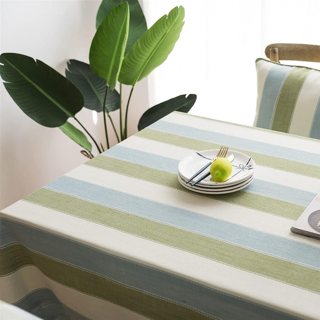 A table with a blue and green striped tablecloth features a stack of white plates with a green apple and cutlery on top. A potted plant with large green leaves is in the background near a window.