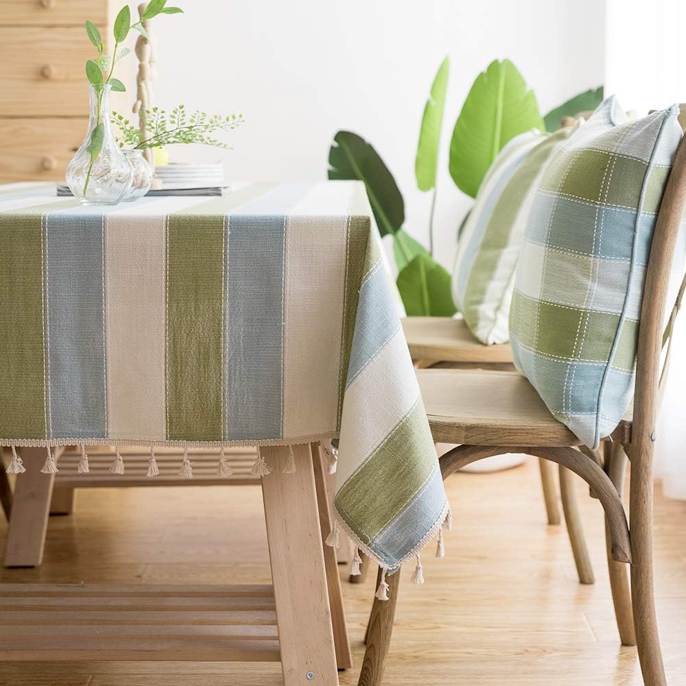 A cozy dining area with a wooden table covered by a striped tablecloth in green, blue, and beige. Two wooden chairs with matching plaid cushions are beside the table. A vase with leaves decorates the table, and large green plants are in the background.