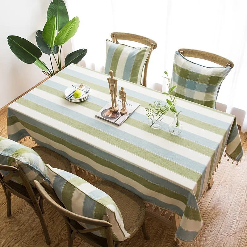 A dining table covered with a green and beige striped tablecloth. Two wooden chairs with plaid cushions are visible. The table is decorated with a small green plant, a bowl of fruit, and a wooden mannequin. A potted plant is nearby on the floor.