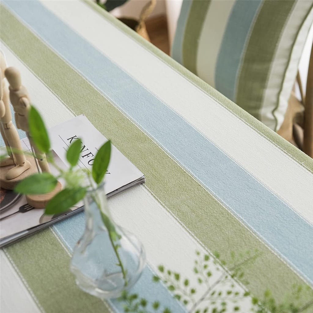 A table with a striped tablecloth in green, blue, and white, featuring a clear vase with leafy stems, a small mannequin hand, and a magazine titled “KIN” partially visible. A striped cushion is in the background.