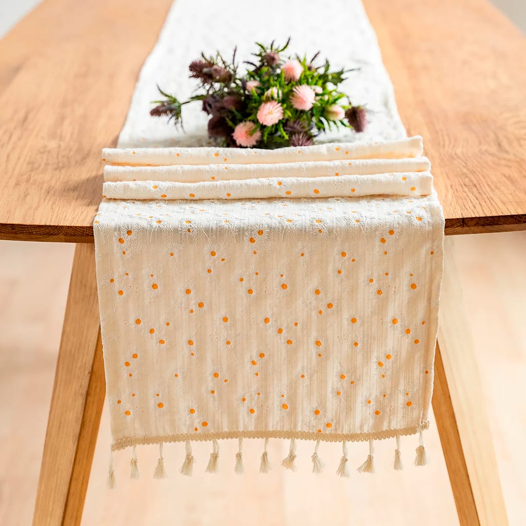 A wooden table is adorned with a cream table runner featuring small orange dots and tassels at the ends. A bouquet of purple and pink flowers is placed on the runner.