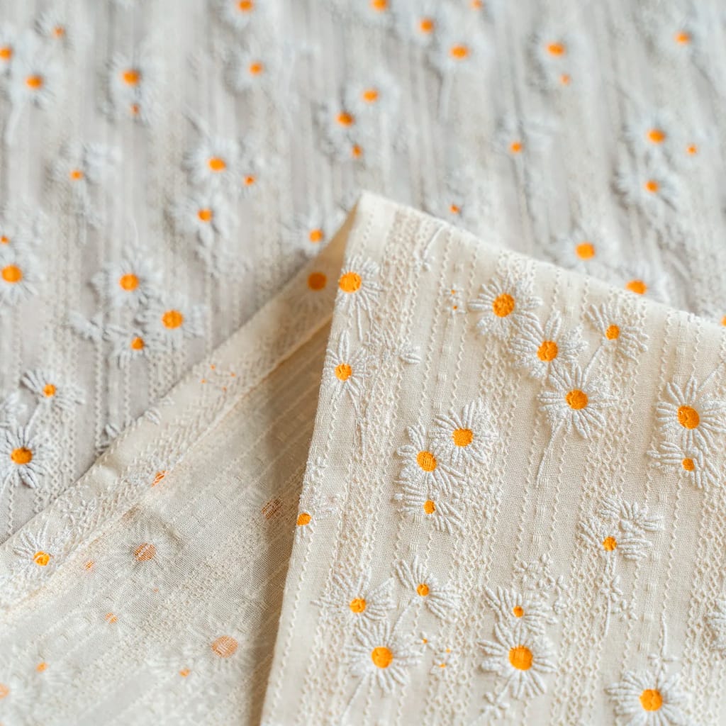 Close-up of an elegant cream-colored fabric featuring an embroidered pattern of white daisies with orange centers. The fabric is gently folded, showcasing its texture and intricate design.