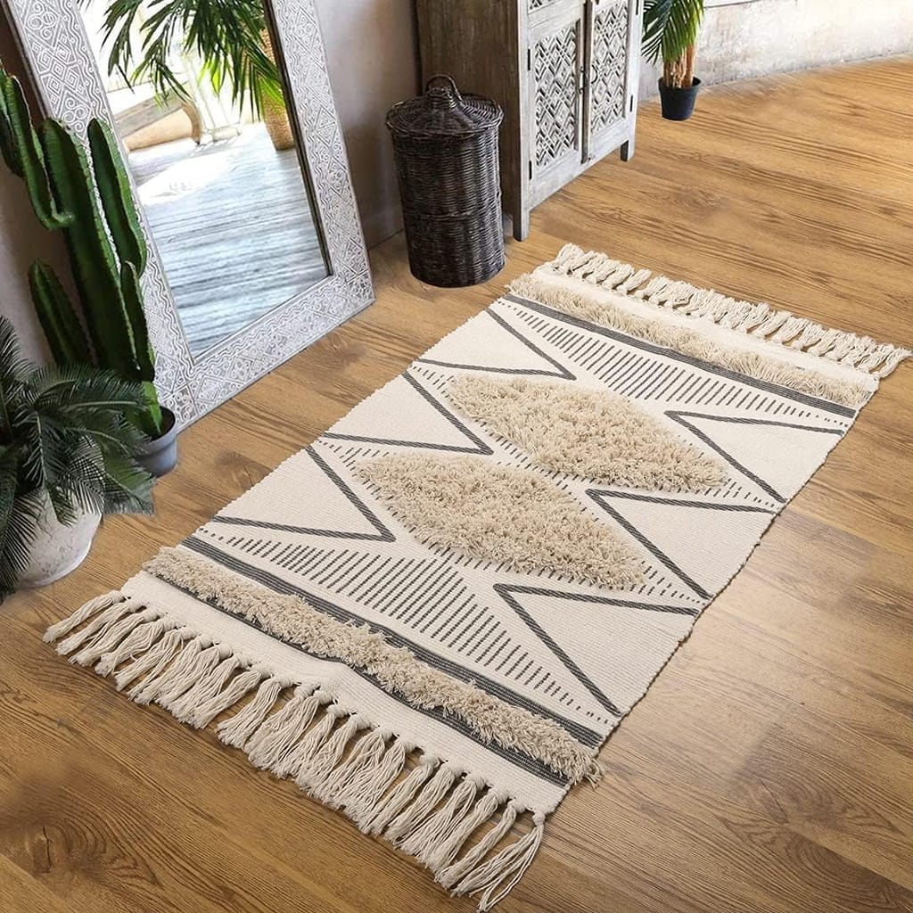 A beige and gray geometric-patterned rug with fringed edges lies on a wooden floor. Surrounding decor includes a large mirror, wicker basket, and various potted plants, enhancing the cozy, bohemian atmosphere.