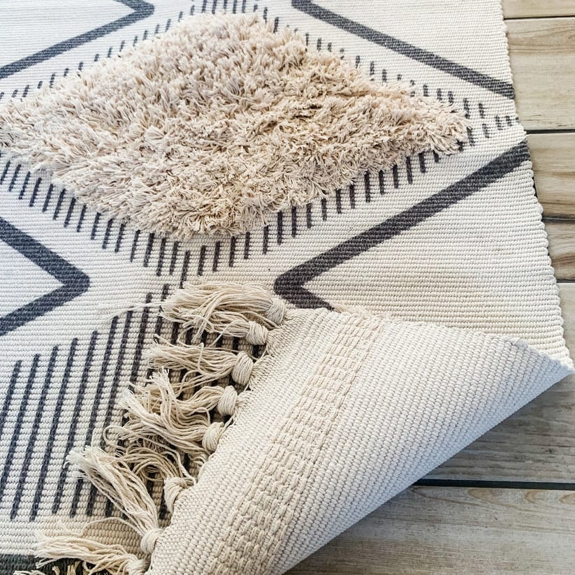 A folded geometric-patterned rug with diamond shapes and fringed edges lies on a wooden floor. The rug features a mix of textures, with raised and flat woven areas in shades of beige and gray.