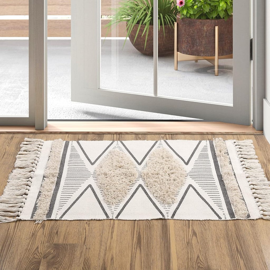 A geometric-patterned rug with tassels on a wooden floor, placed near an open glass door leading outside. A large planter with green foliage and pink flowers is visible outside on the patio.