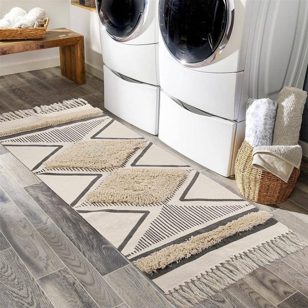 A laundry room with a geometric-patterned rug on a wooden floor. Two front-loading washing machines are stacked. A wicker basket holds folded towels, and a wooden bench is in the background.