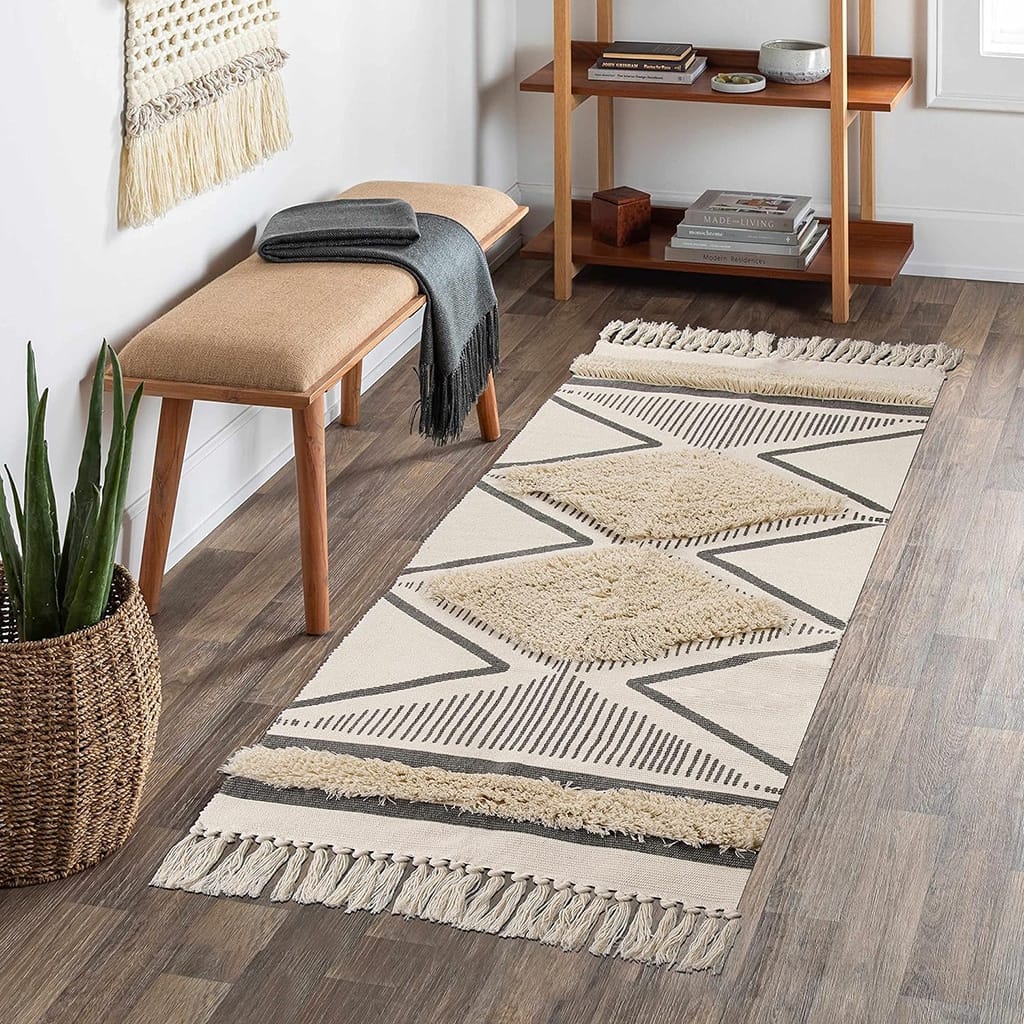 A cozy room features a woven rug with geometric patterns on a wooden floor. A wooden bench with folded blankets sits beside a window. A wooden shelf holds books and decor. A woven basket with a plant adds greenery to the space.