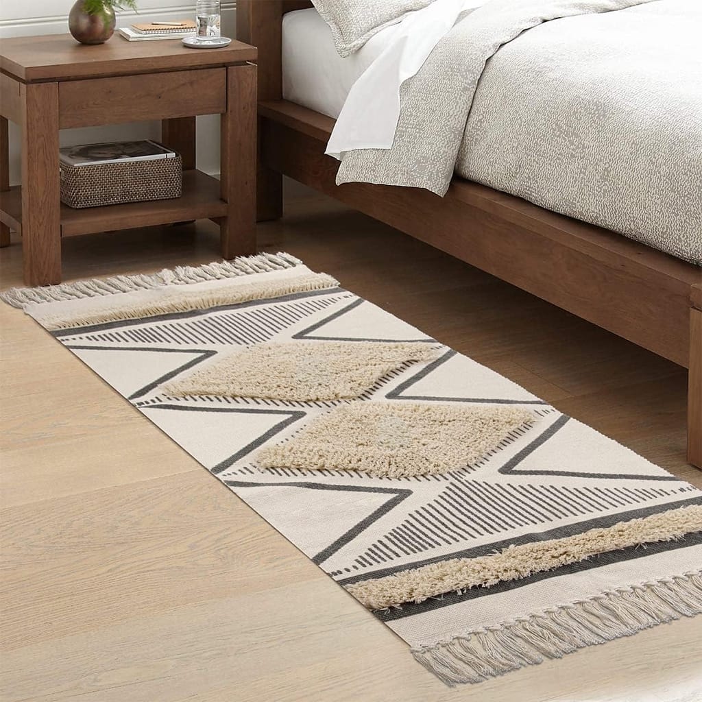 A beige and brown geometric pattern rug with tassels is placed beside a wooden bed in a bedroom. A wooden nightstand with a plant and a woven basket is next to the bed on a light wooden floor.