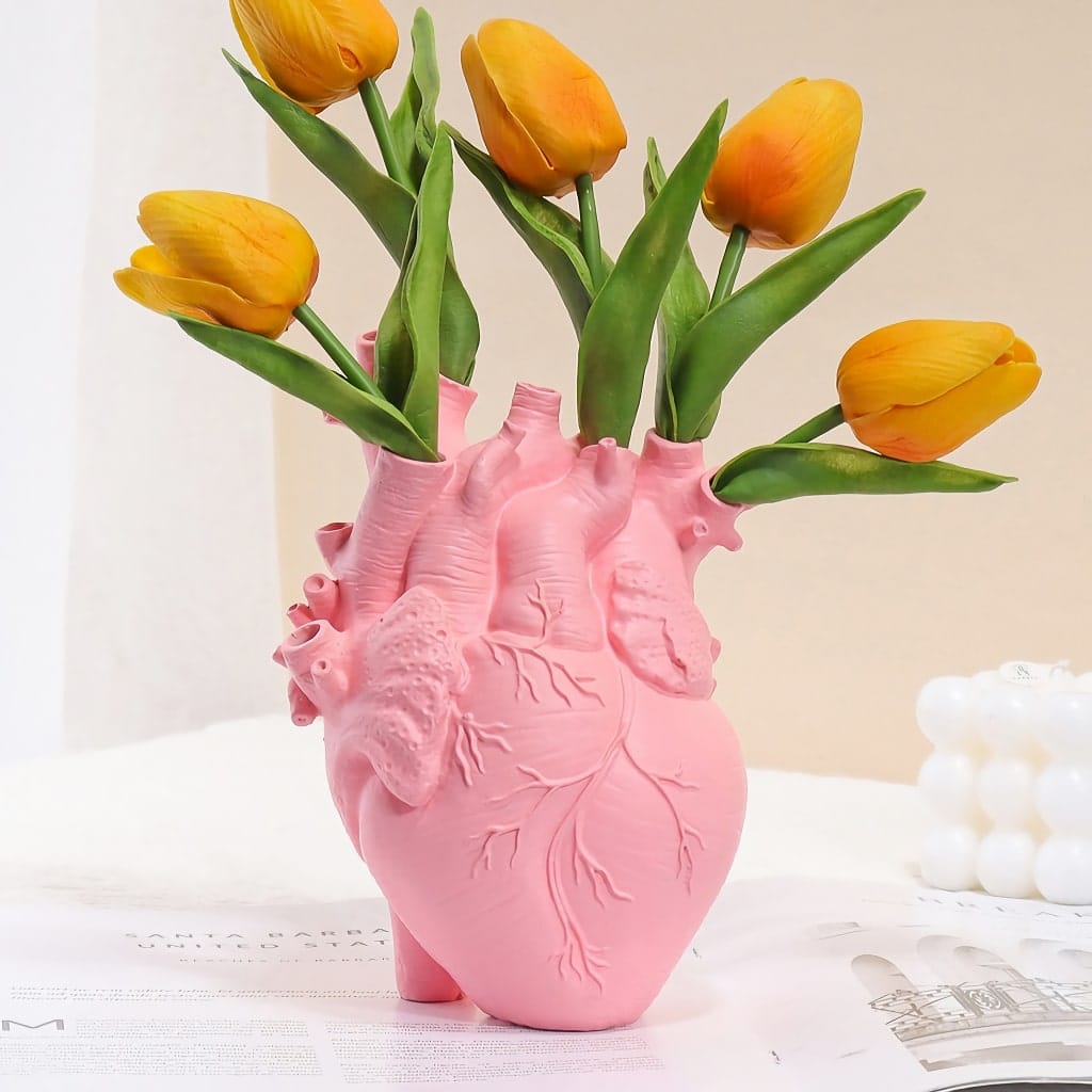 A pink vase shaped like an anatomical heart, with yellow tulips blooming from the top, is placed on a light surface. A blurred magazine or paper is partially visible in the background.