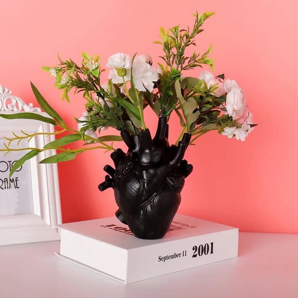 A black vase shaped like a human heart holds flowers with white blossoms and green leaves. The vase sits on a white book with the date September 11, 2001 printed on it. The background is a coral pink wall.