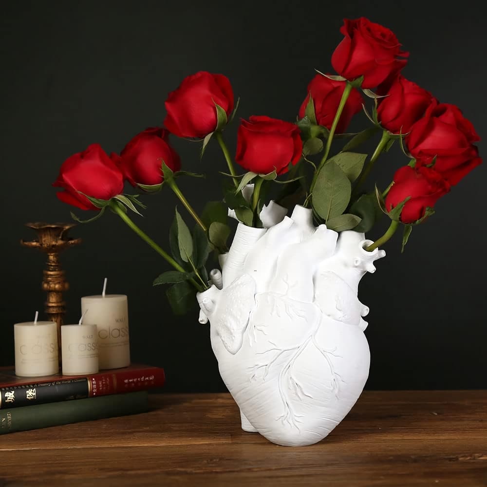 A white vase shaped like a human heart holds nine vibrant red roses with green stems and leaves. The vase is placed on a wooden table, alongside stacked books and lit candles, against a dark background.