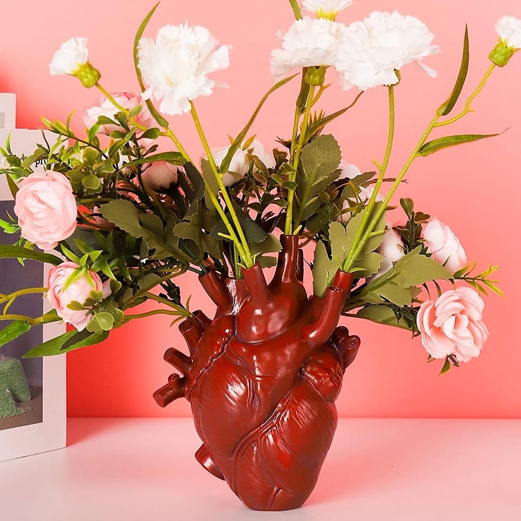 A vase shaped like a human heart holds an arrangement of white and pink flowers. The background is a soft pink, enhancing the vibrant colors of the floral display and the unique design of the vase.