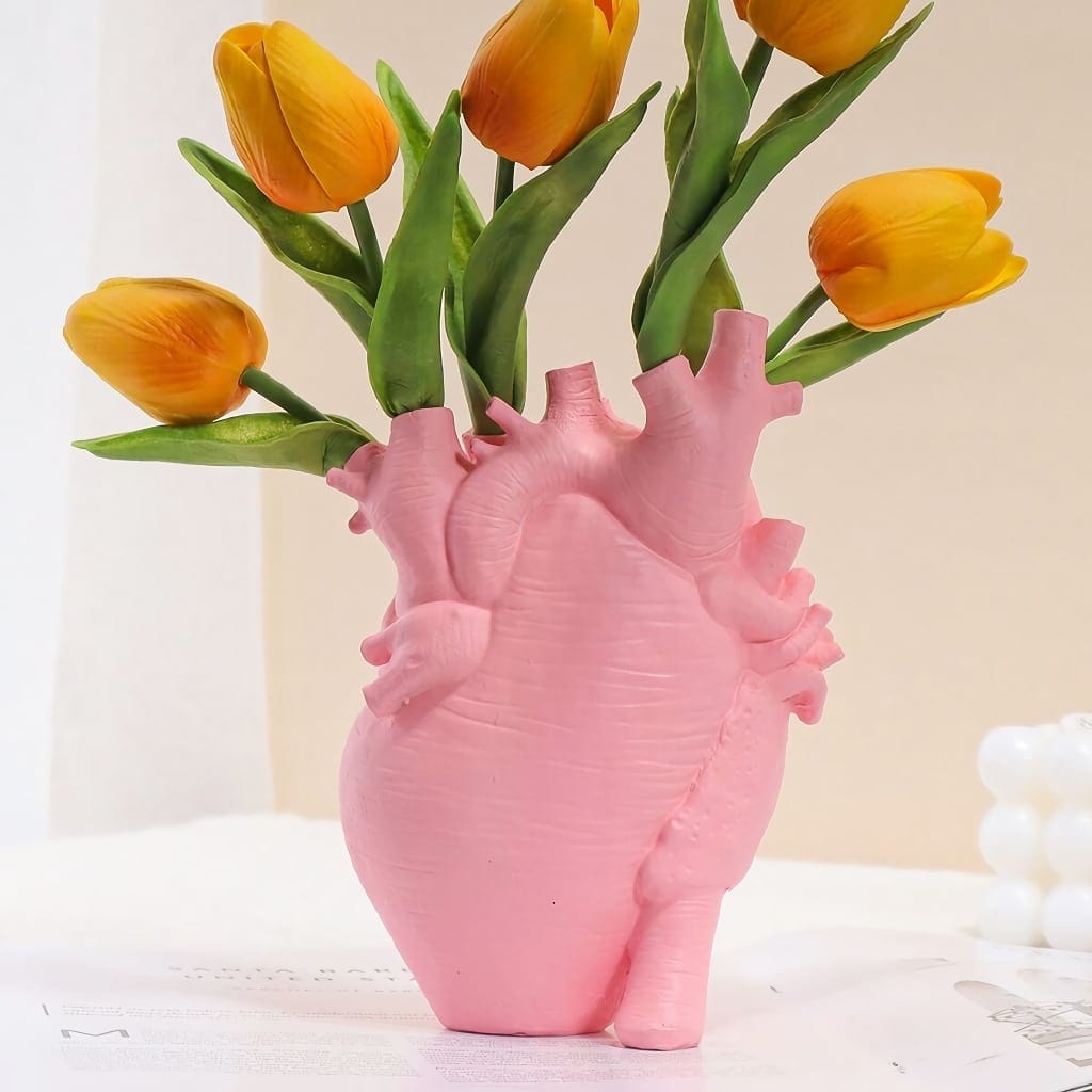 A unique vase shaped like a pink human heart holds several yellow tulips with green leaves. The background is softly lit, creating a delicate and artistic atmosphere.