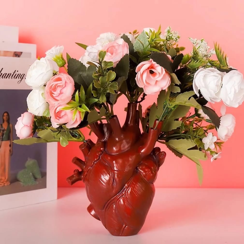 A heart-shaped vase holds a bouquet of pink and white roses with greenery against a pink background. A blurred magazine is visible on the left.