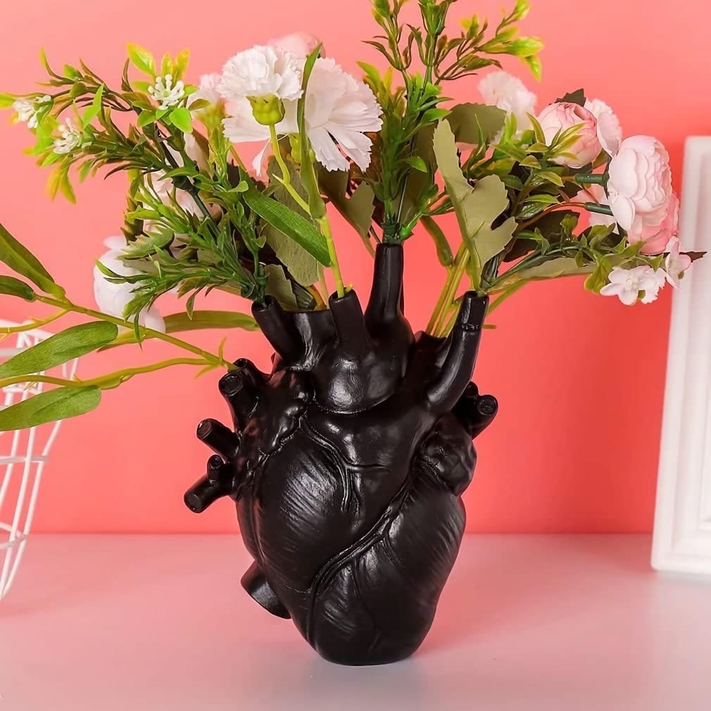 A black vase shaped like a human heart holds a bouquet of white and pink flowers with green leaves. The background is a vivid coral color, and the vase sits on a light pink surface.