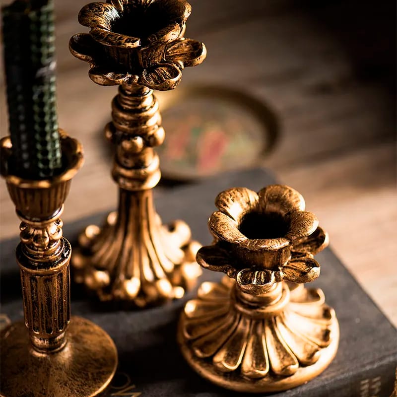 Close-up of three ornate brass candle holders with floral designs on a wooden surface. The candle holders have a vintage look, and one has a dark, twisted candle placed in it. The background is softly blurred.