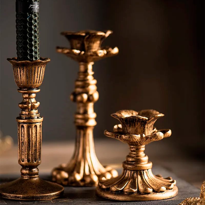 Three ornate, gold-toned candlestick holders of varying heights are displayed. They have intricate detailing with fluted and floral designs, enhancing their vintage appeal. One holder contains a partially visible black candle.