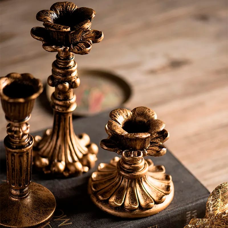 Antique gold candle holders with intricate floral designs are placed on a wooden surface. They feature different heights and ornate bases, capturing a vintage and elegant aesthetic.