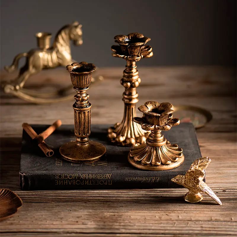 A set of three ornate, gold candlesticks of varying heights are placed on an old book atop a wooden surface. A small gold bird figurine and a cinnamon stick are nearby. Blurred in the background is a brass rocking horse.