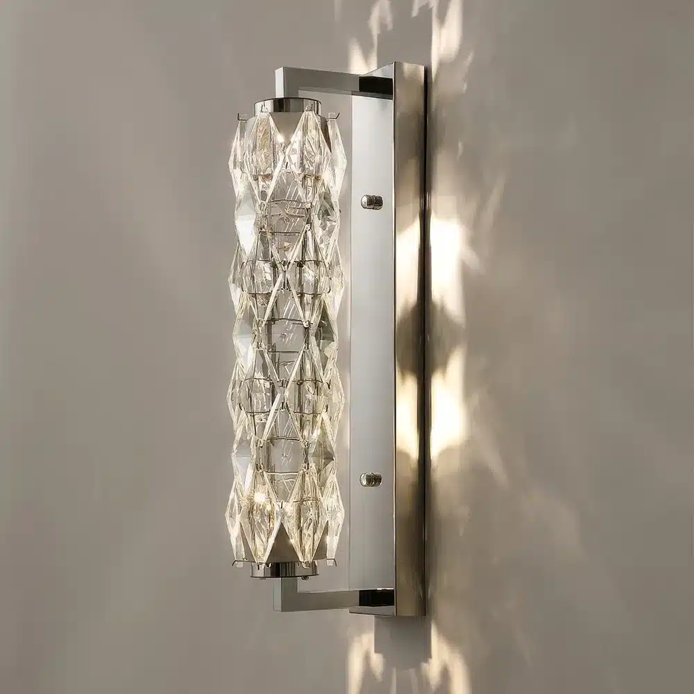 A modern wall sconce with a rectangular metal base and a cylindrical glass design. The glass has a diamond pattern that creates a sparkling light effect on the surrounding wall. The fixture is mounted vertically.